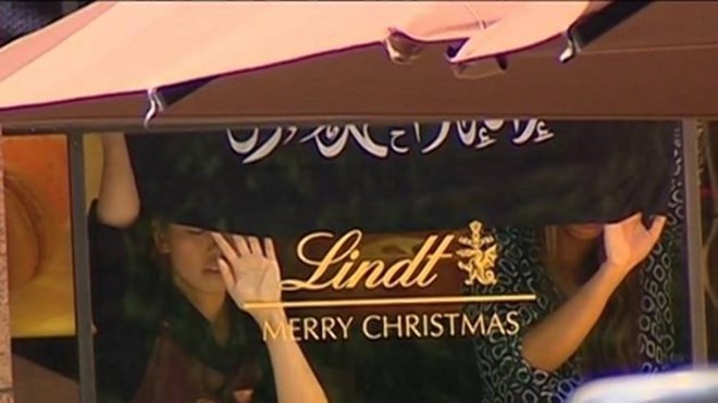 Các con tin đứng bên trong quán Lindt Chocolat Cafe với 2 tay đặt lên cửa sổ, dưới lá cờ đen có chữ Arab.